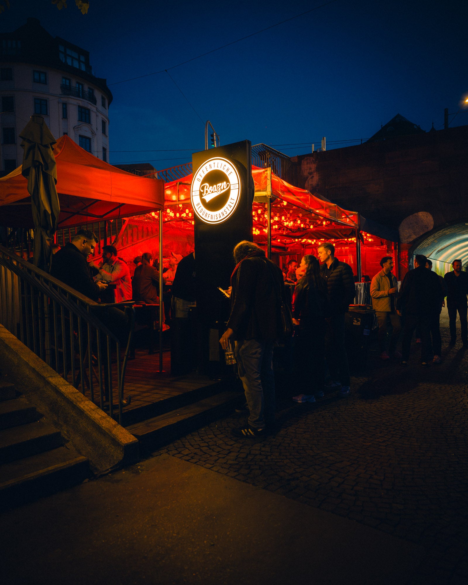 Boazn-Pavillon mit beleuchtetem Schild Öffentliche Bedürfnisanstalt bei Dämmerung an der Ludwigsbrücke – Boazn München, Bar an der Isar, Mai 2026