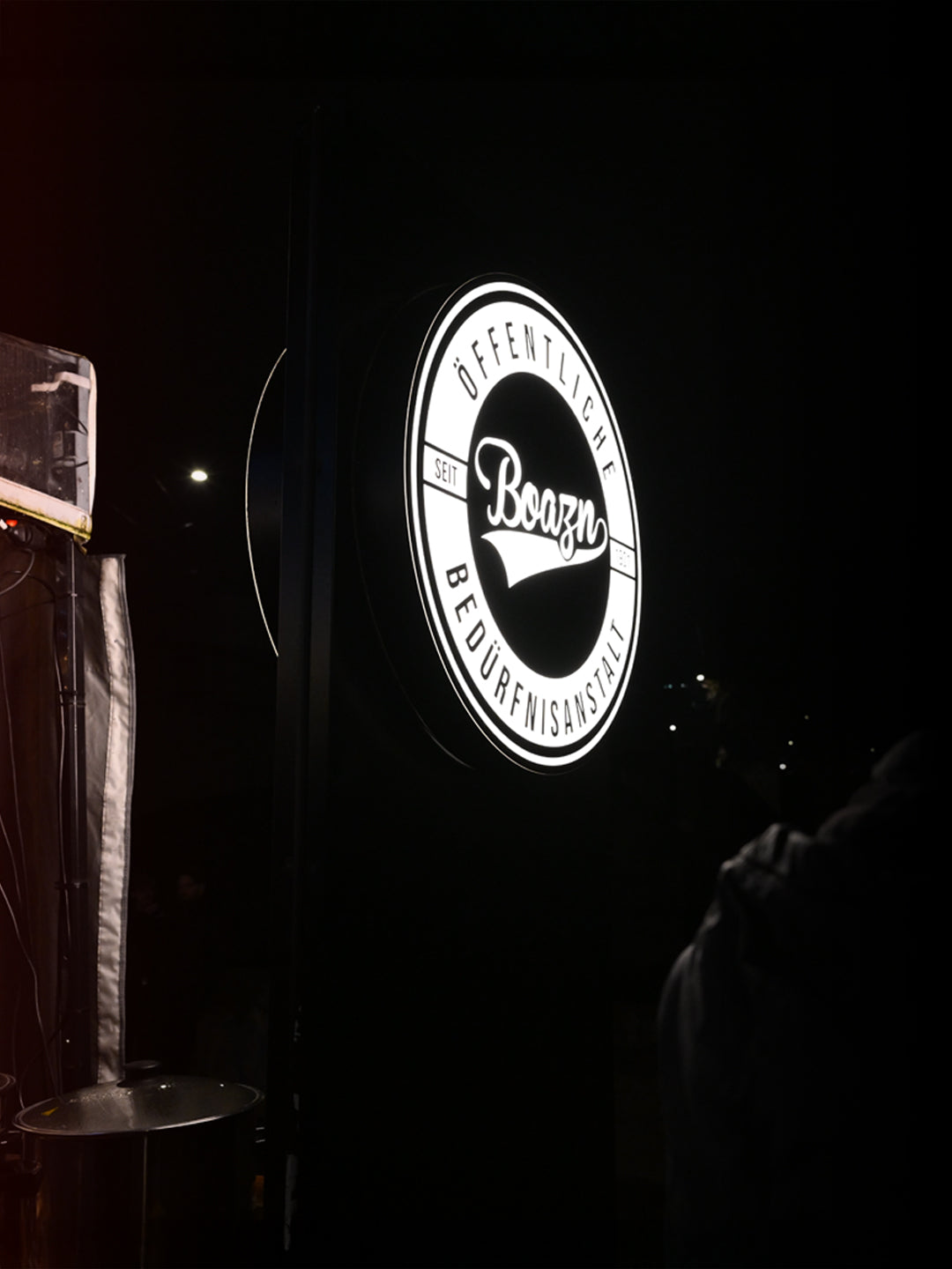 Beleuchtetes Boazn-Logo-Schild „Öffentliche Bedürfnisanstalt seit Boazn“ leuchtet im Dunkel der Nacht – Bar an der Ludwigsbrücke, München, Januar 2026