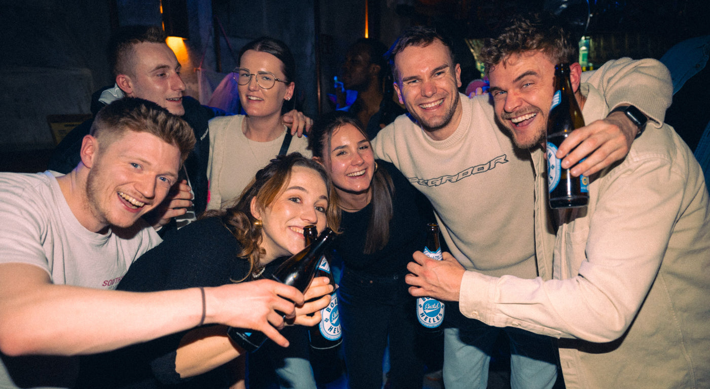 Sechs Freunde lachen und stoßen mit Bristol-Helles-Flaschen an – Bar an der Ludwigsbrücke, März 2026 8