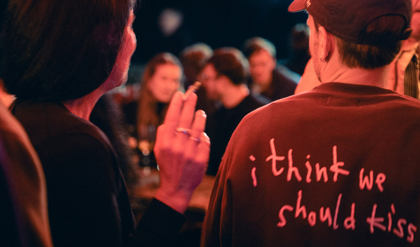 Rückseite eines roten Sweatshirts „i think we should kiss“ neben Frau mit erhobener Zigarette – Bar an der Ludwigsbrücke, Februar 2026 12