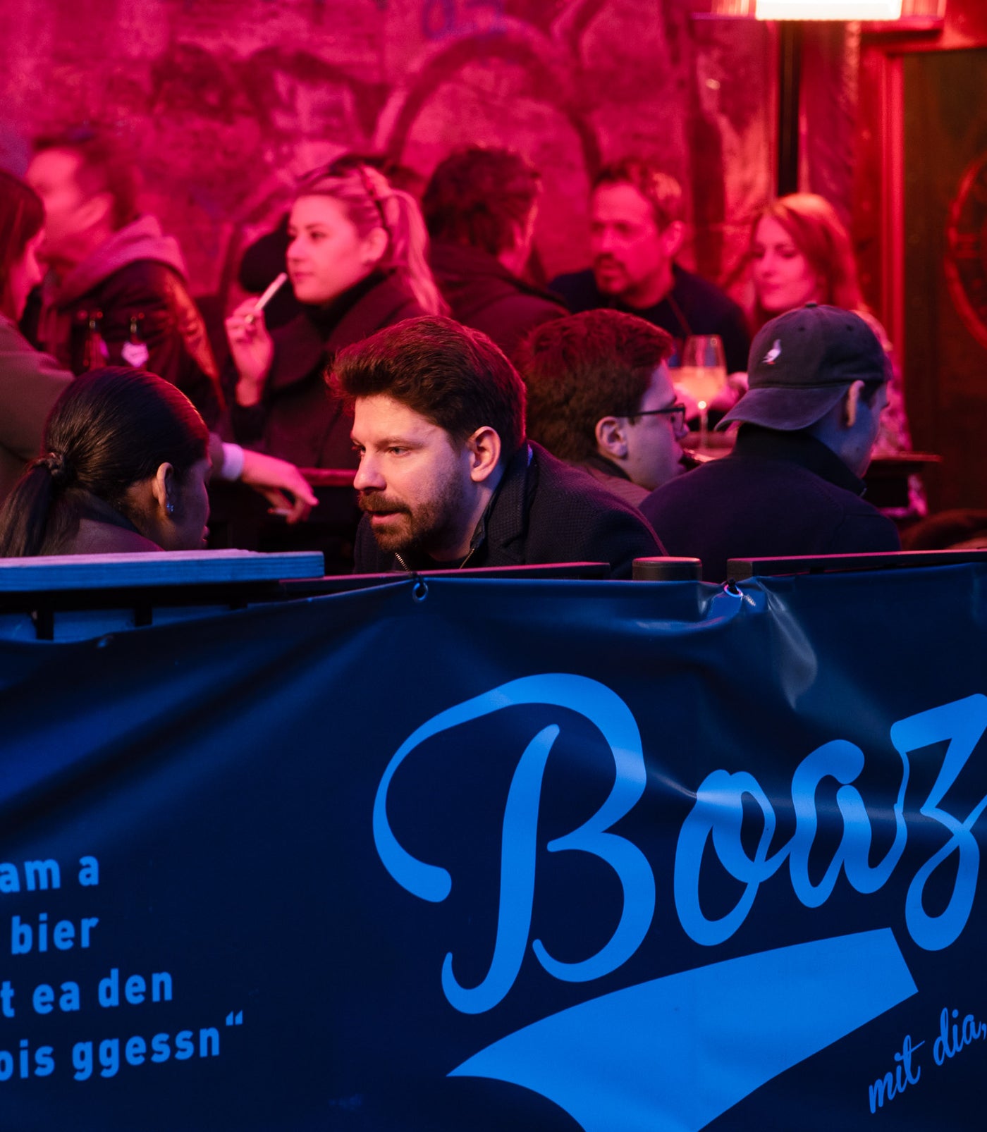 Bartender und Gäste hinter „Am a Bier mit dia“-Boazn-Banner unter rotblauem Licht – Kneipe in München, Januar 2026 5