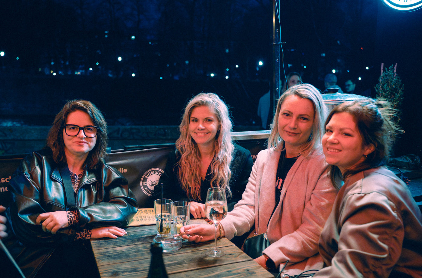 Vier Frauen am Holztisch der Boazn-Terrasse mit Weißwein, Lichter der Isar im Hintergrund – Bar an der Ludwigsbrücke, Februar 2026 15