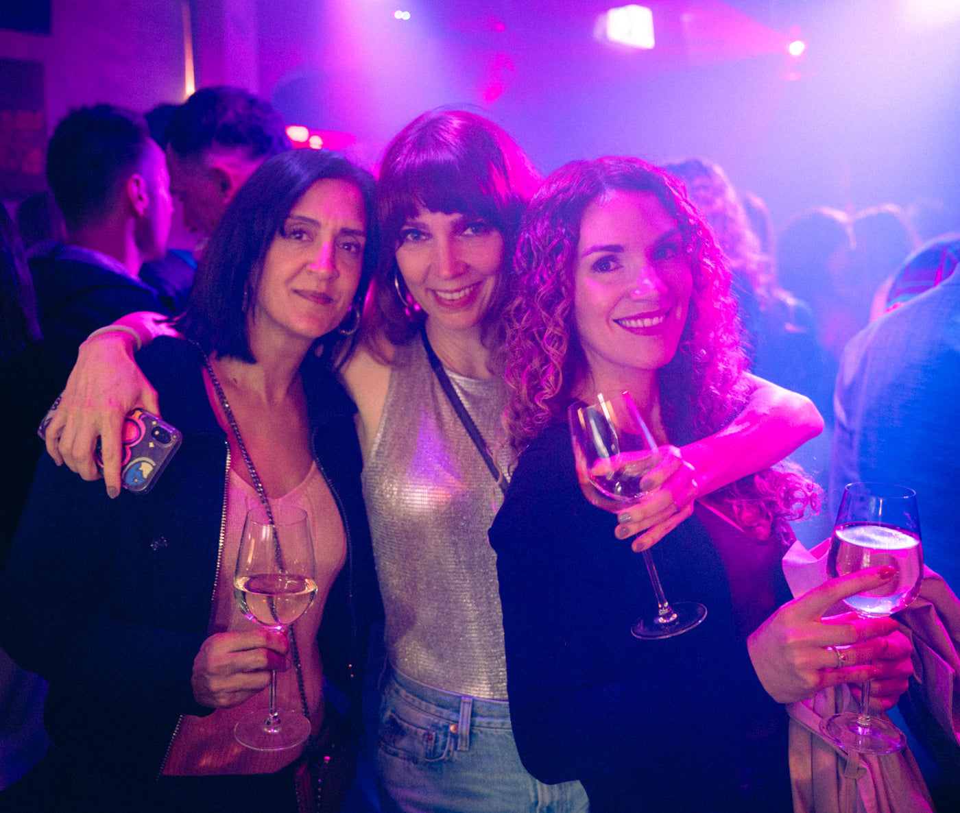 Drei Freundinnen umarmen sich lachend mit Sektgläsern unter pinkem Discolicht – Tanzbar in München, Februar 2026 16