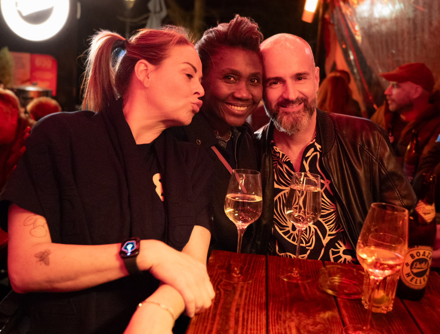 Drei Freunde umarmen sich, Frau in der Mitte mit Kussmund, daneben Mann im Hemd mit Weißwein – Bar in München, März 2026 10