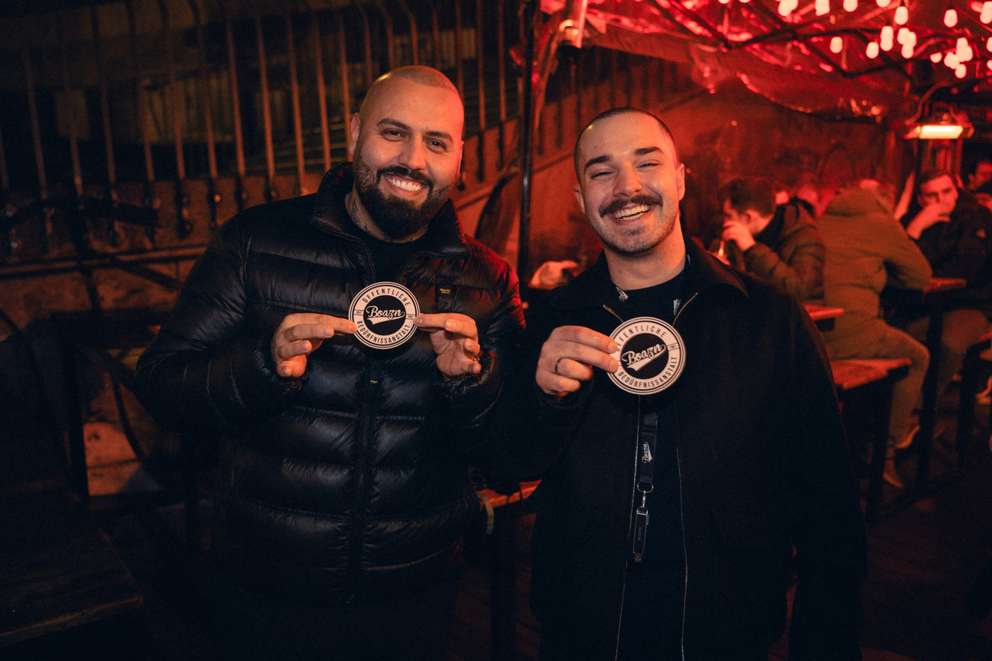 Zwei Männer halten Boazn-Bierdeckel mit „Öffentliche Bedürfnisanstalt“-Logo hoch – Kneipe in München, Februar 2026 21