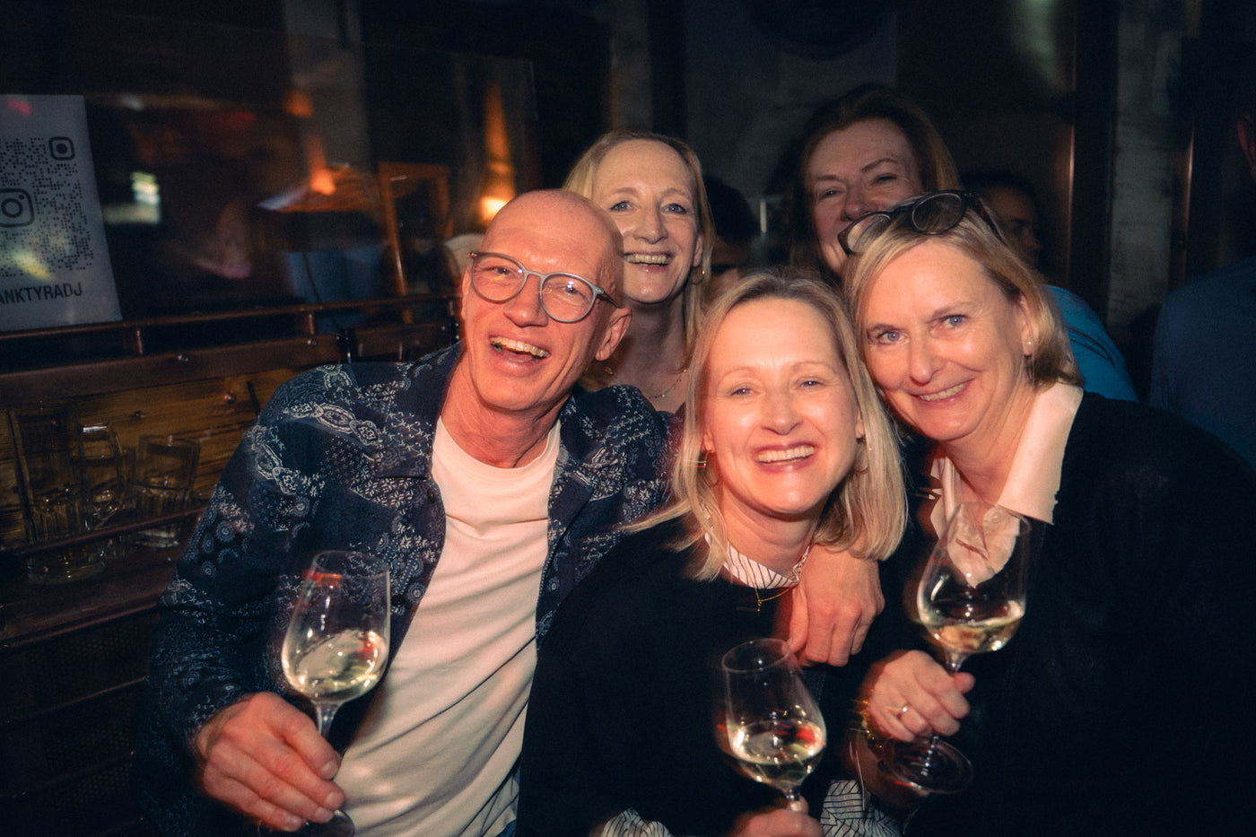 Fünf Erwachsene umarmen sich mit Weißweingläsern vor dem QR-Code-Schild von Franky Tyra DJ – Bar an der Ludwigsbrücke, Februar 2026 25