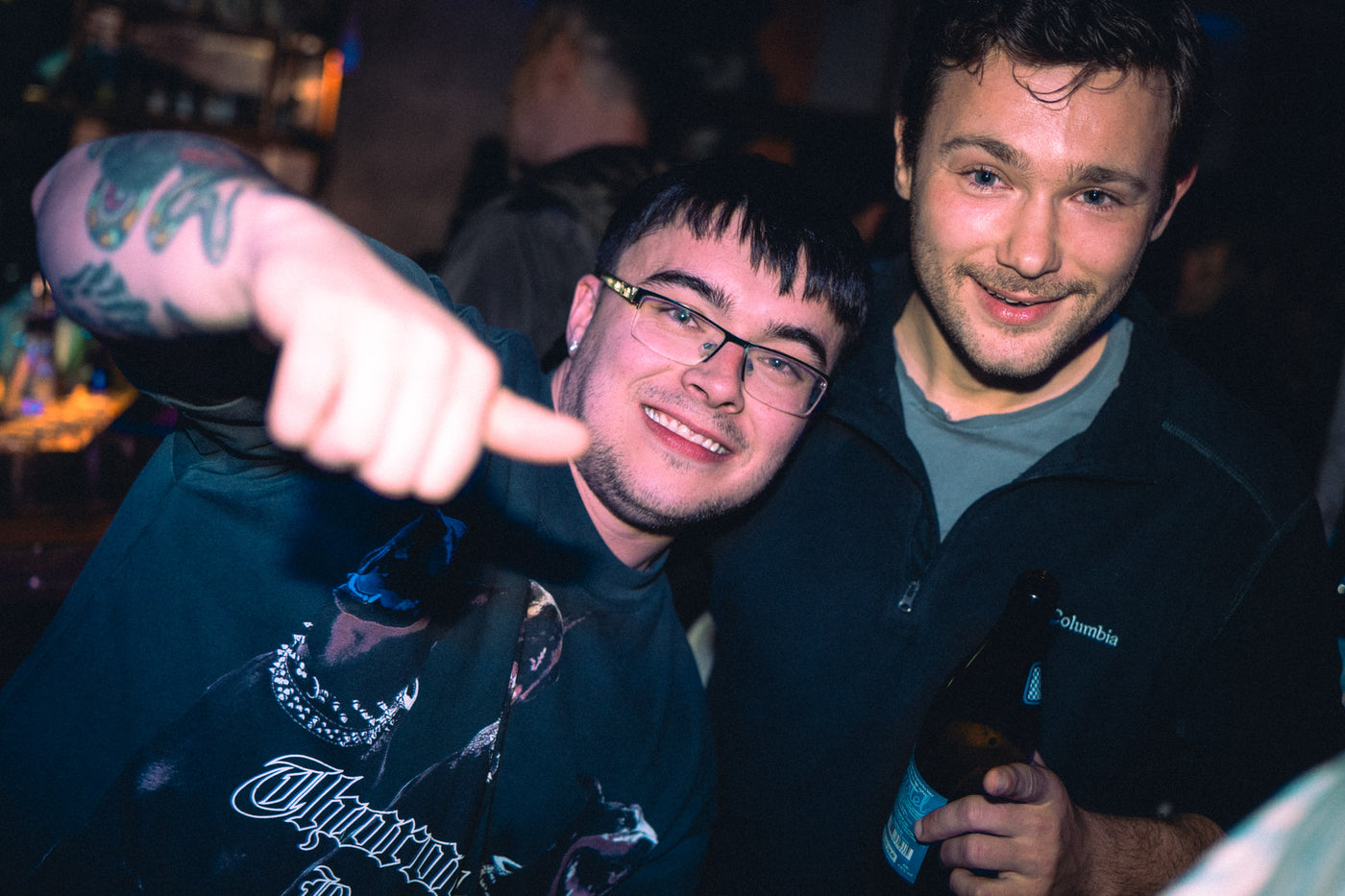 Zwei Freunde, einer mit Tattoo-Arm und Brille zeigt zur Kamera, der andere mit Augustiner-Bier – Kneipe in München, März 2026 5