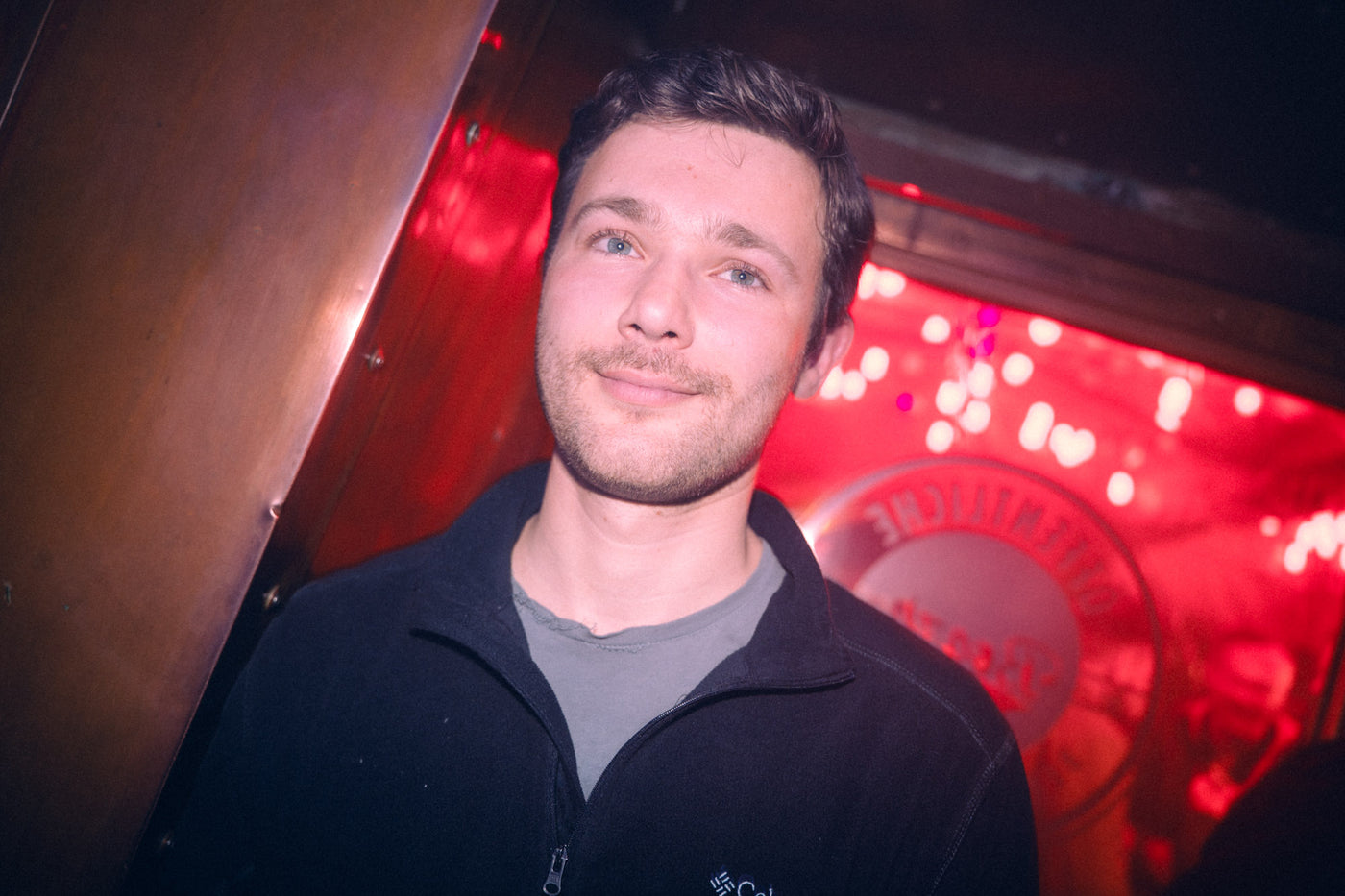 Junger Mann lächelt unter rotem Licht vor dem Boazn-Wandschild „Öffentliche“ – Bar in München, Januar 2026 12