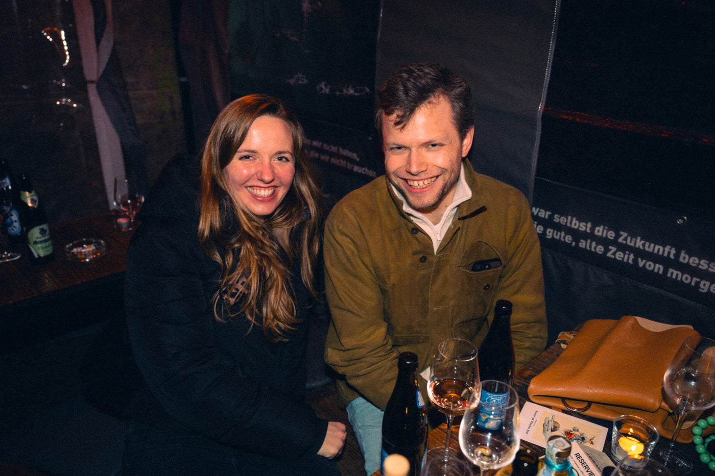 Paar lacht am Tisch mit Augustiner-Flasche und Weingläsern vor Boazn-Wandtext – Bar in München, Januar 2026 15