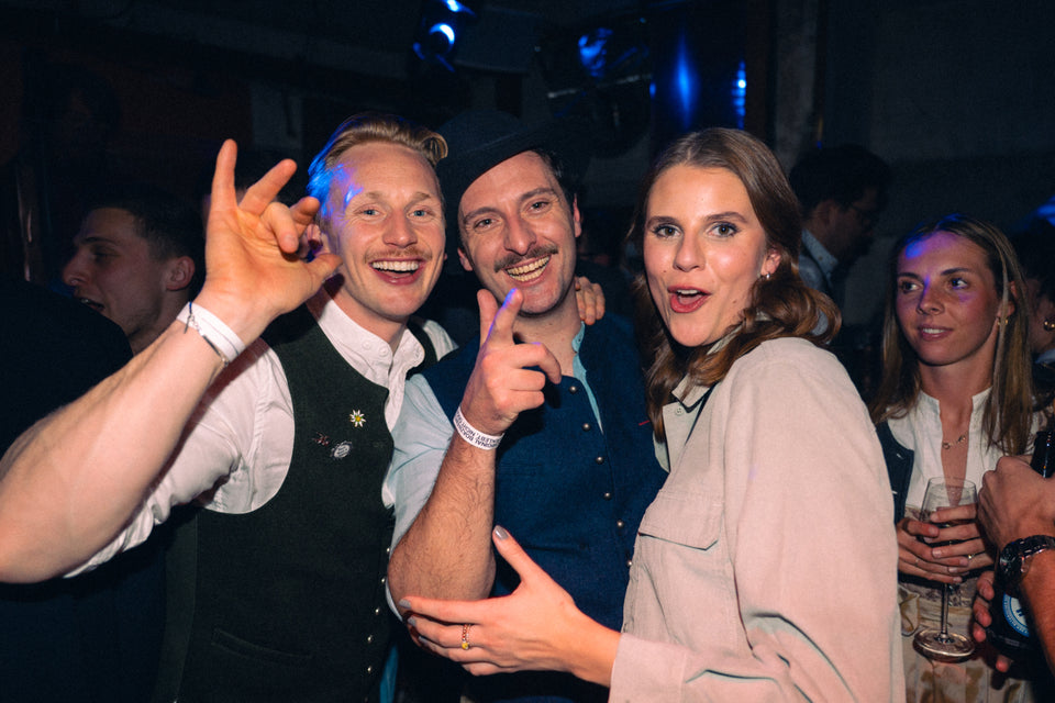 Drei Freunde in Trachtenwesten und mit Hut lachen ausgelassen, eine Frau zeigt zur Kamera – Nightlife München, Februar 2026