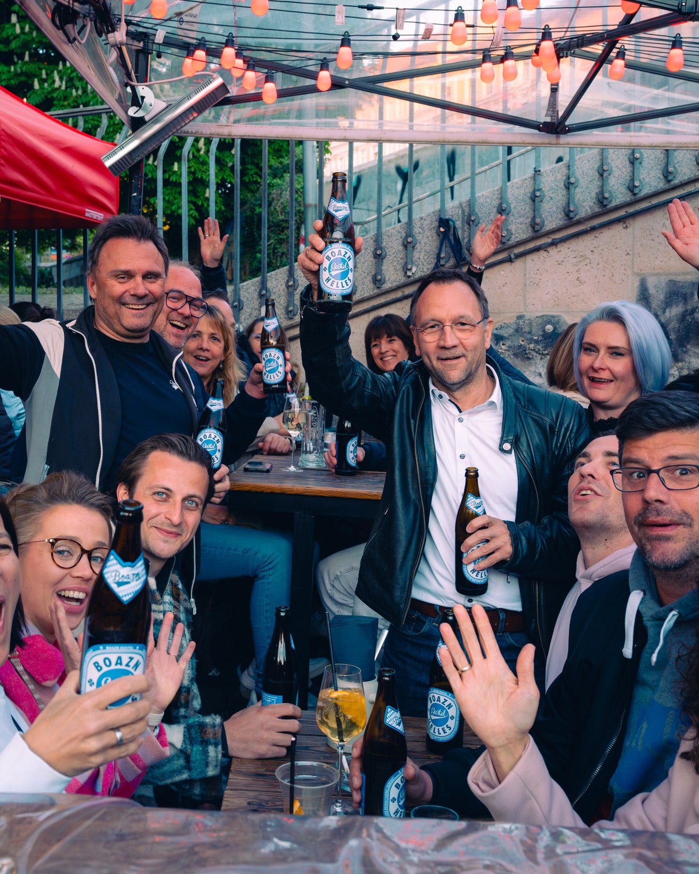 Gruppe stößt mit Boazn-Eigenbier auf der Terrasse unter der Ludwigsbrücke an – Boazn München, April 2026