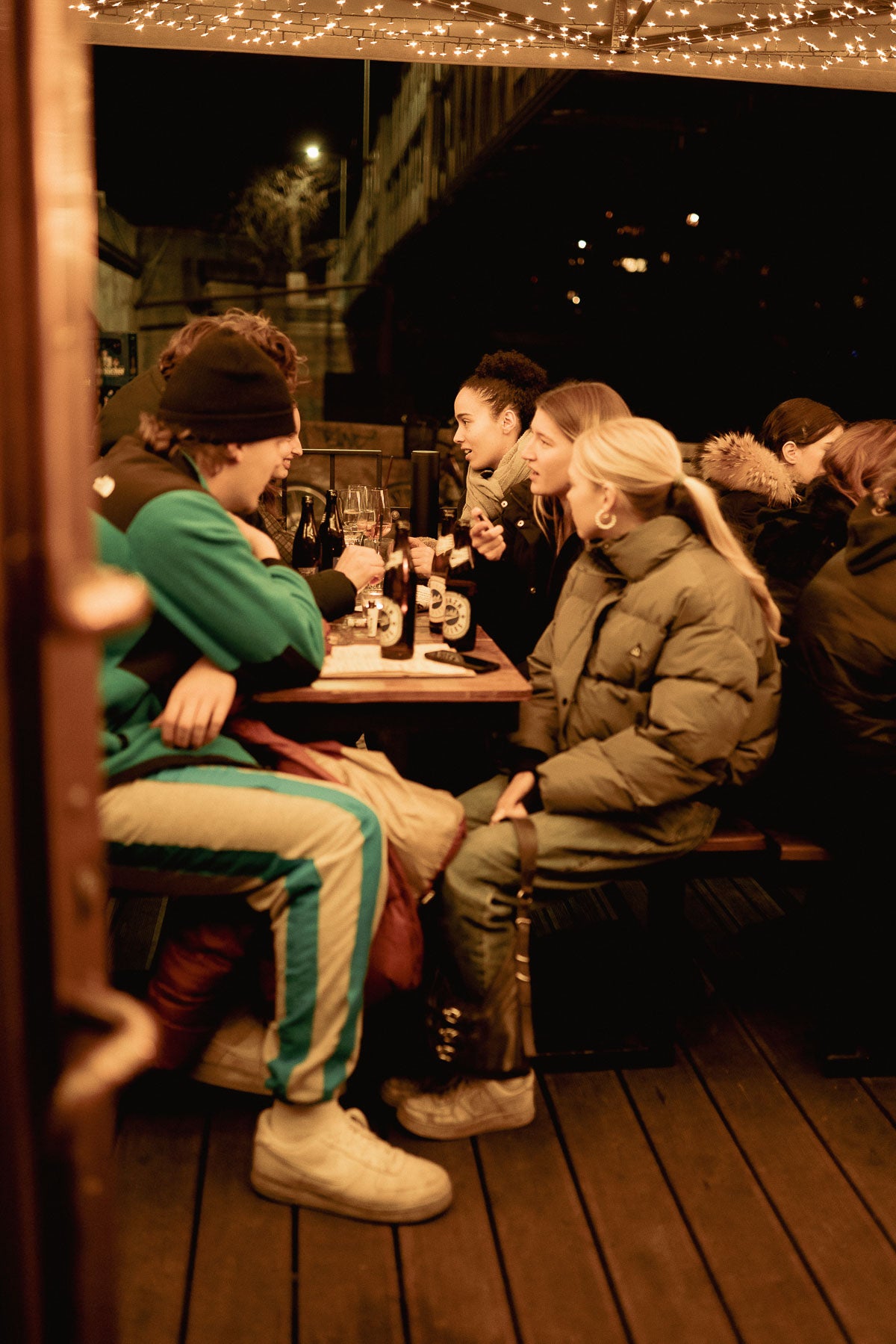 Freundesgruppe am Tisch auf der Terrasse mit Wein und Boazn Helles – Bar an der Isar, München, Dezember 2025 6
