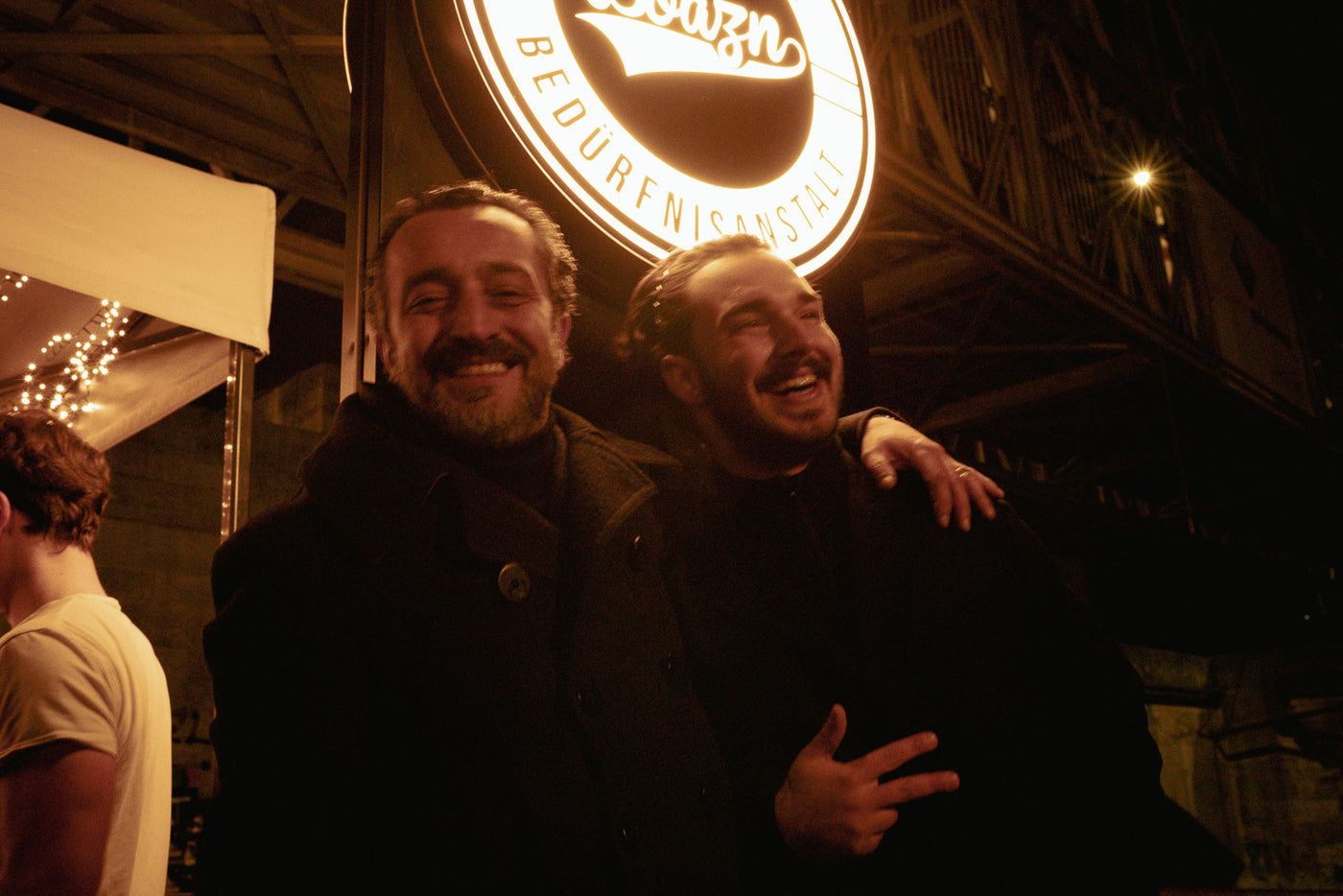 Zwei Freunde lachen ausgelassen vor dem leuchtenden Boazn-Logo – Bar an der Ludwigsbrücke, München, Dezember 2025 24