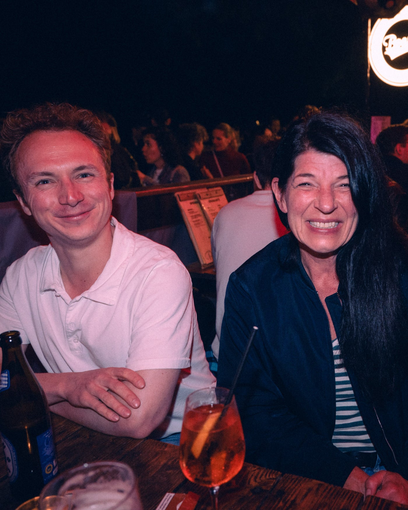 Junges Paar lacht am Holztisch, Bierflasche und Aperol Spritz im Vordergrund – Boazn München, Tanzbar an der Ludwigsbrücke, Mai 2026 3