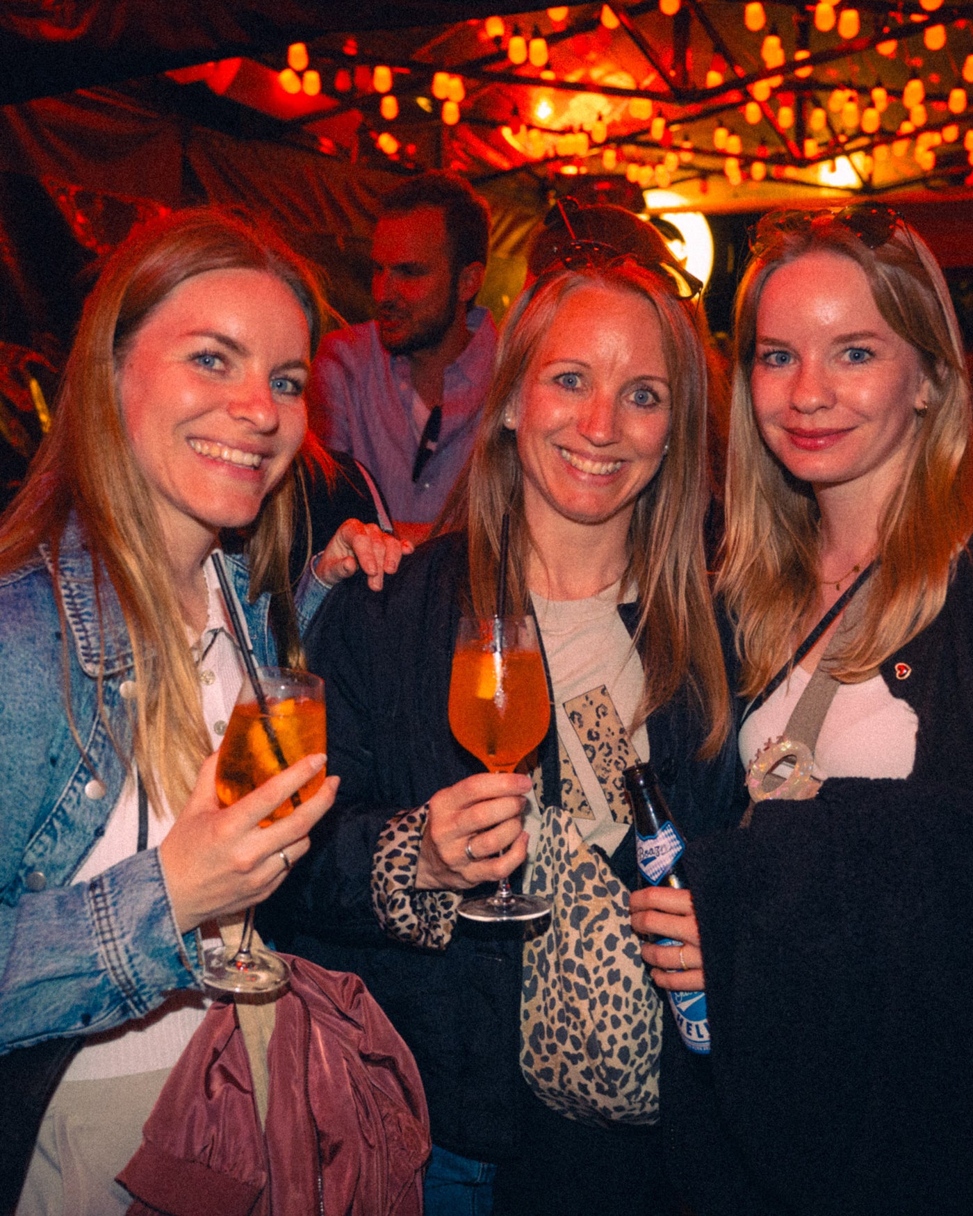 Drei Frauen strahlen mit Aperol Spritz unter den warmen Lichterketten – Boazn München, Nightlife an der Isar, Mai 2026 9