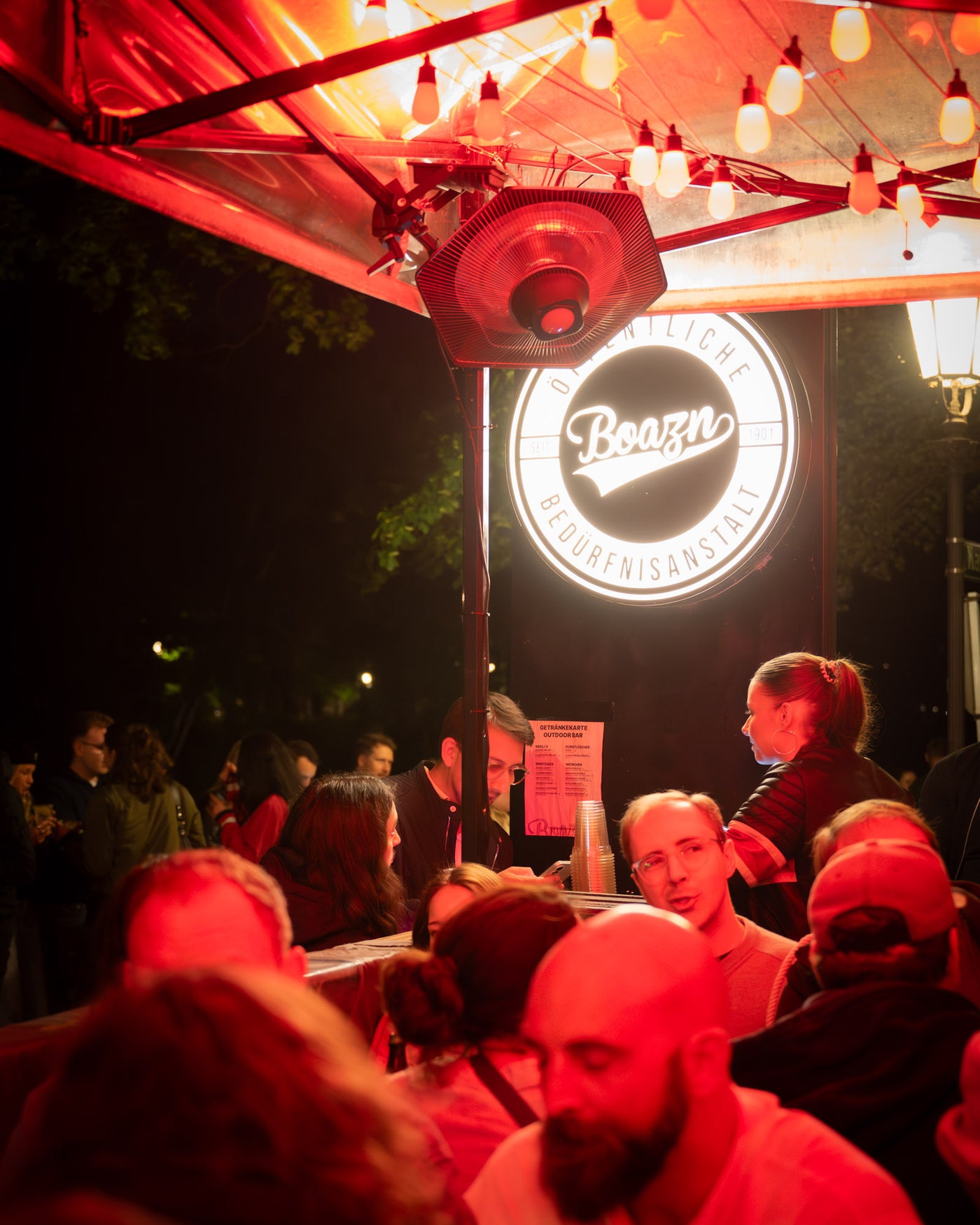 Voller Outdoor-Tresen unter Heizpilz und Boazn-Leuchtschild bei Nacht – Boazn München, Bar an der Isar, Mai 2026 21