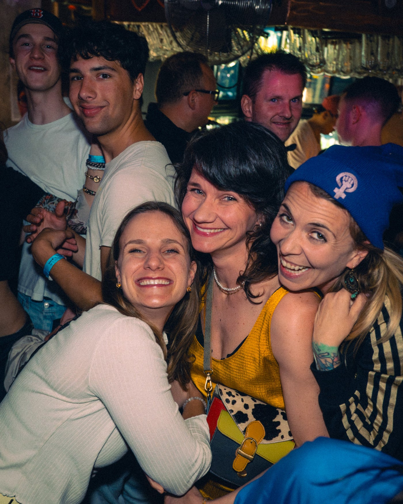 Drei Freundinnen umarmen sich vor der alten Theke, zwei Freunde lachen dahinter – Boazn München, Bar an der Ludwigsbrücke, Mai 2026 26
