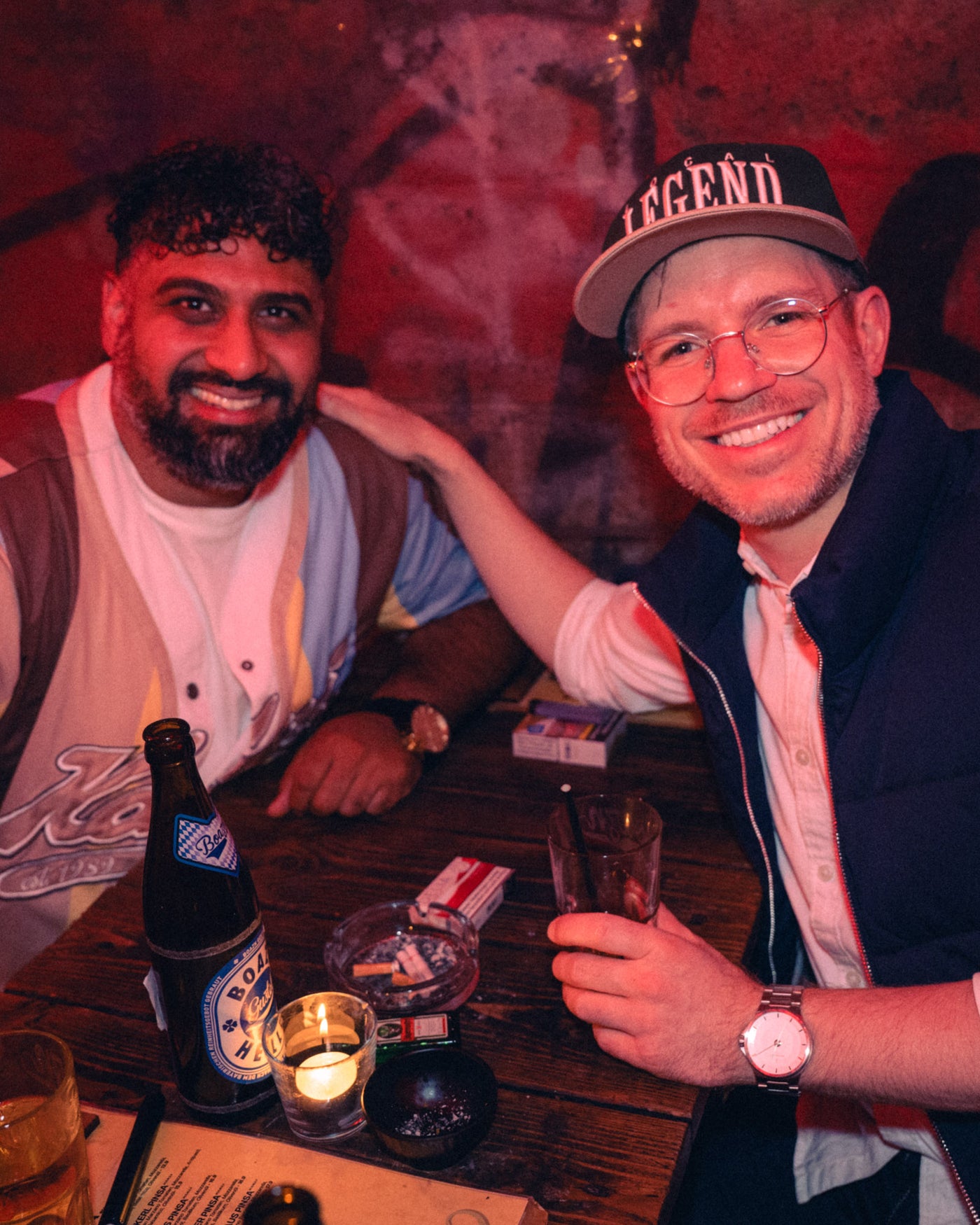Zwei Freunde lachen am Tisch mit Augustiner und Kerze im Vordergrund – Boazn München, Bar an der Ludwigsbrücke, Mai 2026 36