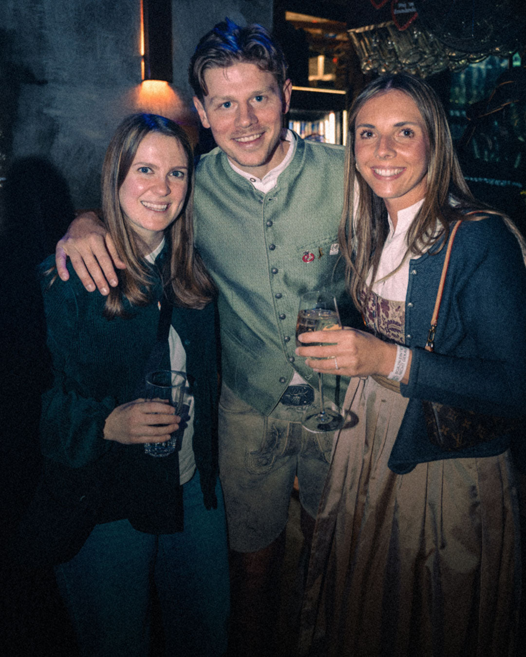 Mann mit Lederhose und blau gefärbter Tolle umarmt zwei Frauen in Tracht – Tanzbar an der Isar, Februar 2026 10